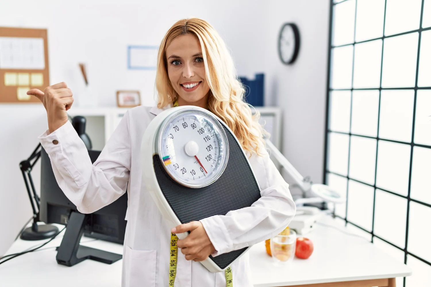 beautiful blonde nutritionist woman holding weight machine balance weight loss pointing thumb up side smiling happy with open mouth